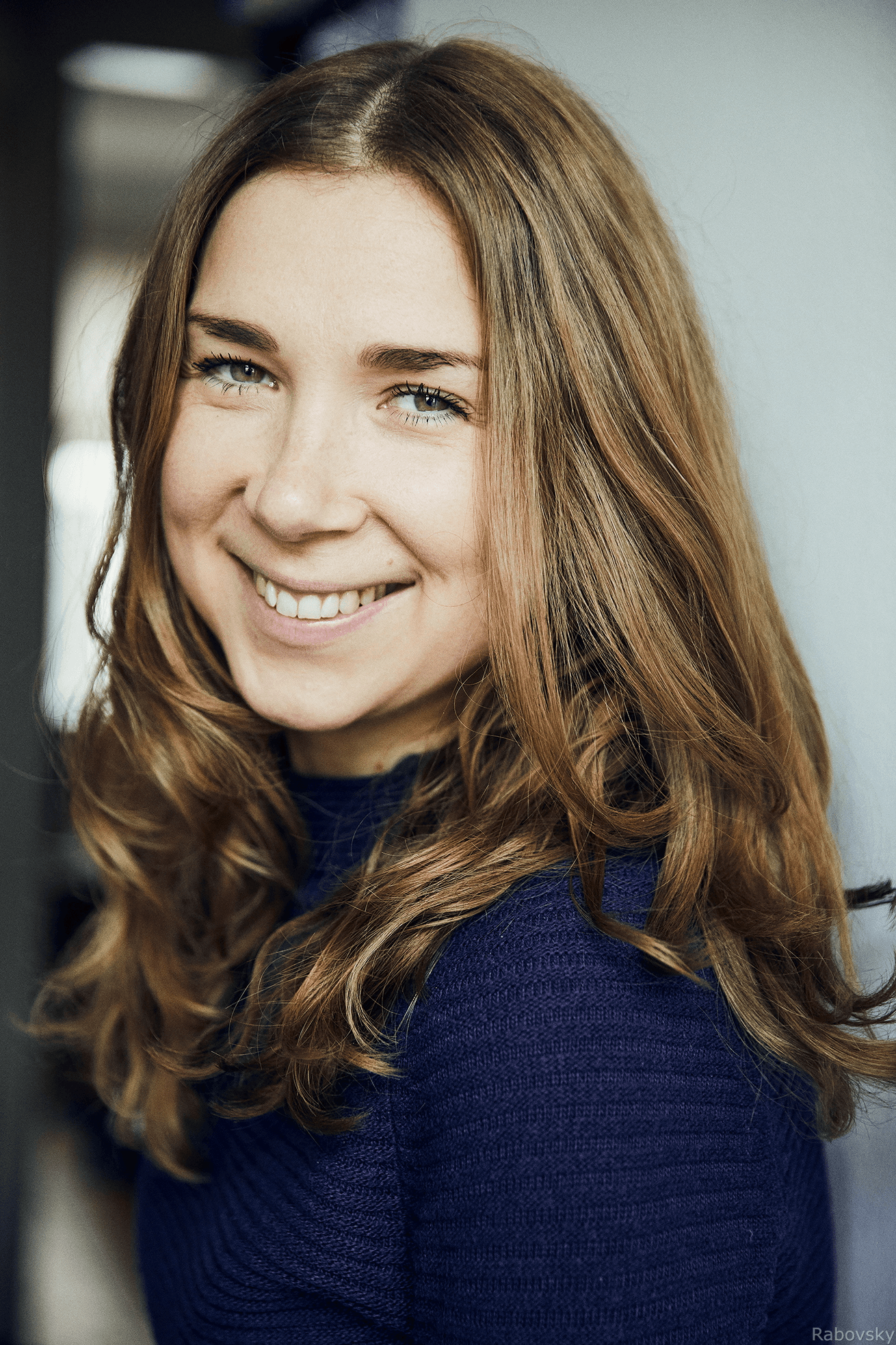 Lächelnde junge Frau mit langen braunen Haaren und blauem Rollkragenpullover, Portrait vor hellem Hintergrund.