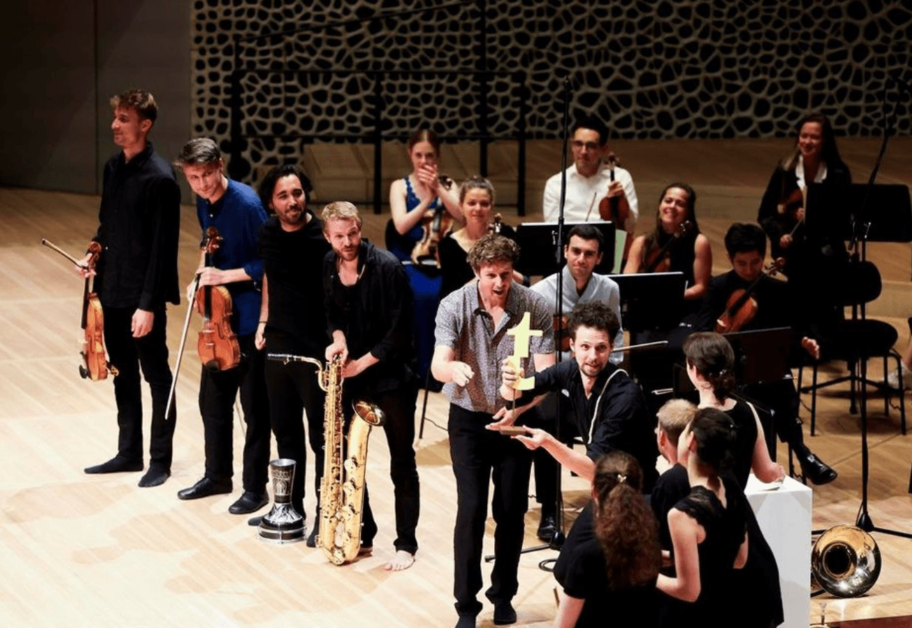Musiker auf einer Konzertbühne; ein Mann hält eine goldene Trophäe, andere halten Geigen und Saxofon, Orchester im Hintergrund.