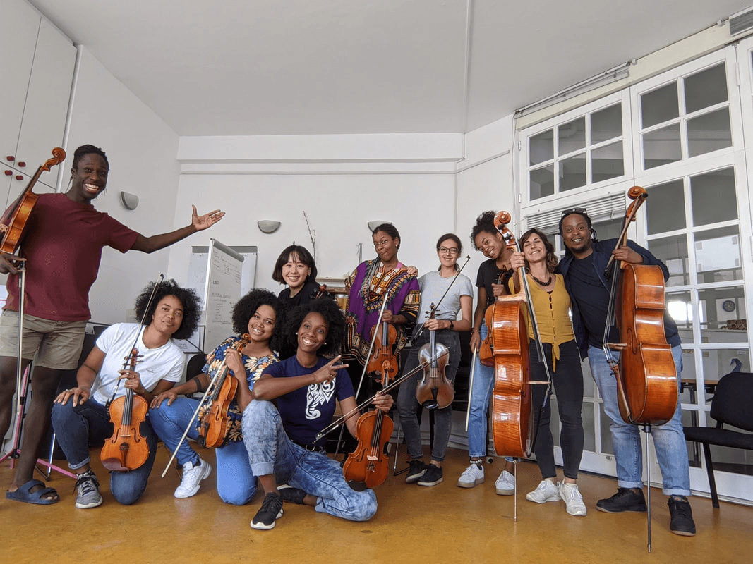 Gruppe von zehn Musikerinnen und Musikern mit Geigen und Celli posiert lächelnd in einem hellen Probenraum.