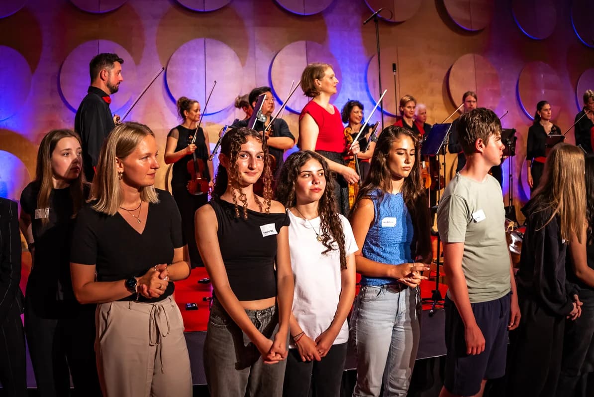 Jugendliche und Erwachsene stehen auf einer Bühne, dahinter Orchestermusikerinnen mit Geigen bei einem Konzert.